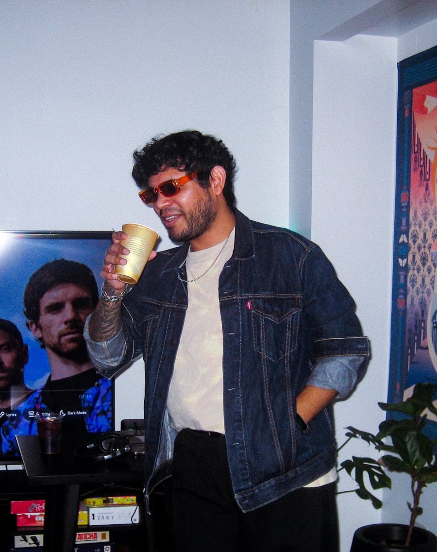 Daniel Campos Lozada con lentes naranjas y campera de jean, tomando de un vaso dorado de plástico en un cumpleaños, con una tele y una planta detrás.